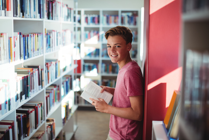 Photographie d'un adolescent souriant tenant un livre dans une bibliothèque.