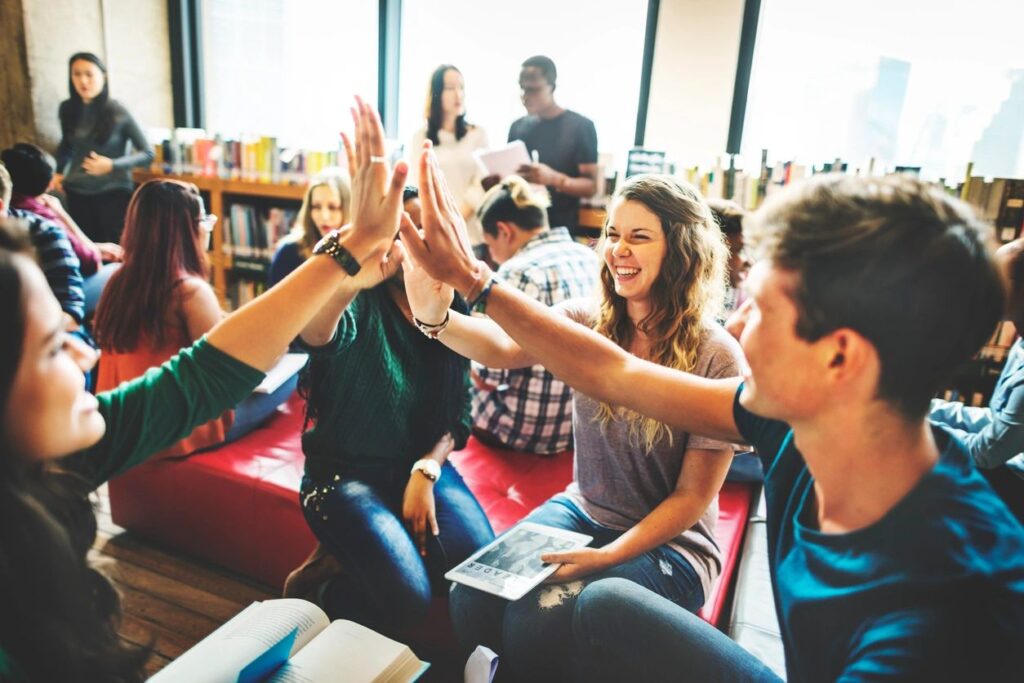 Photographie de quatre adolescents souriants assis en rond dans une bibliothèque, se tapant dans les mains au centre du cercle.