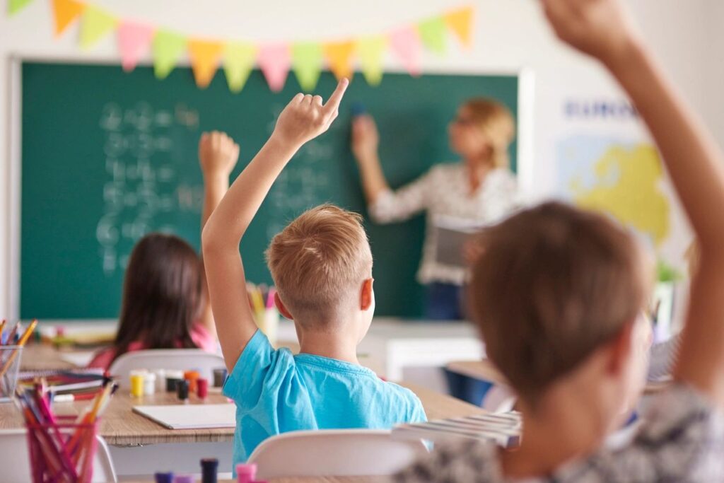 Photographie de trois enfants, de dos et un en arrière de l'autre, levant la main dans leur classe lors d'une leçon de mathématique.