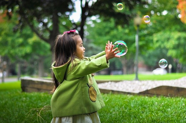 Photographie d'une jeune enfant dans un parc essayant de prendre dans ses mains une grosse bulle en l'air.