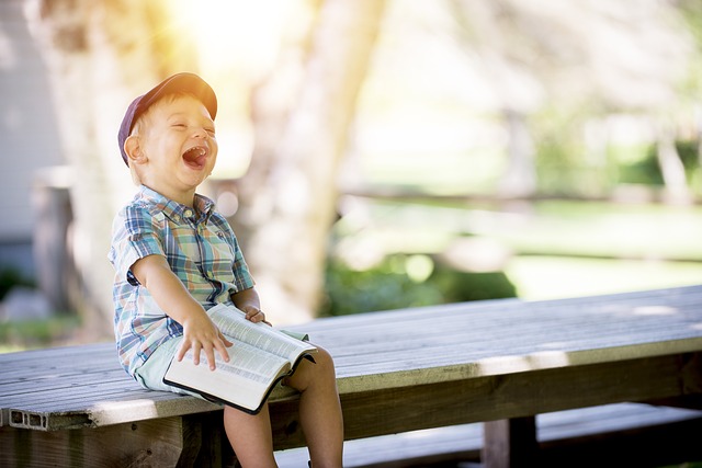 Photographie d'un jeune garçon riant sur une table à pique-nique à l'extérieur, un livre ouvert sur les genoux.